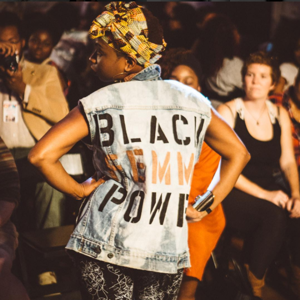 Runway model at Queer Fashion Week wearing a headwrap and hoop earrings stands with with back to us and hands on hips. Jacket reads “Black Femme Power.” The model is being photographed and observed by a crowd of people.