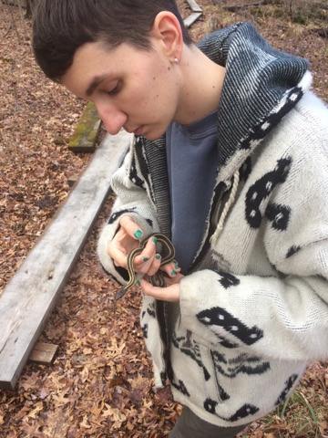 Alt-text: Cade wearing fuzzy zipped sweatshirt, standing outside, with green nail polish, and holding a snake.