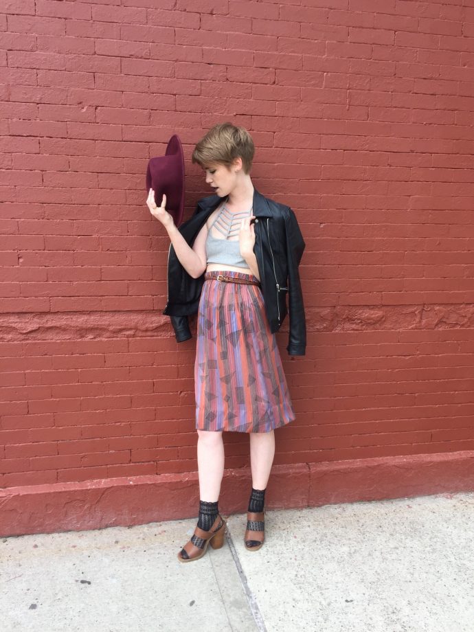 Corrine standing in front of red brick wall with cropped grey top, skirt, brown-heeled sandals, leather jacket, and red fedora in hand.