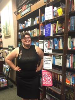 Jaime, wearing a ripped black mesh top and a black pencil skirt, is smiling while standing hands on her hips, in front of a bookshelf of feminist titles at Barnes and Noble.
