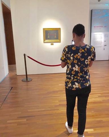 Kayla, a gender non-conforming person of medium height, stands behind a red velvet rope in an art museum, taking a photo of a painting. She is wearing a floral shirt with dark, fitted pants and white slip-on Vans.