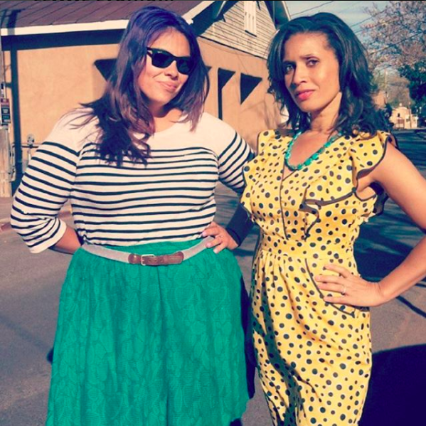  LuzMarina and Anita pose together. LuzMarina is wearing a teal skirt, belt, and striped top; Anita a yellow and black polkadot dress with ruffles along the edges.
