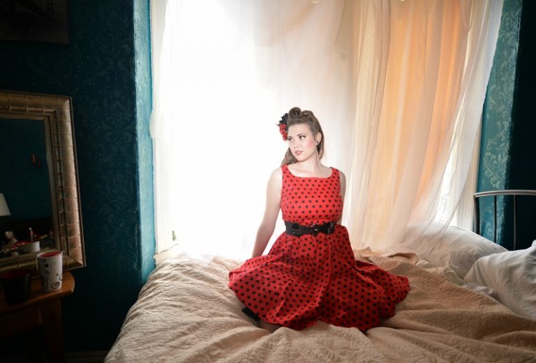 Meghan is sitting in the center of a bed wearing red dress with black polka dots and a red flower in their hair. They are looking off to the side and resting with their shoulders back.